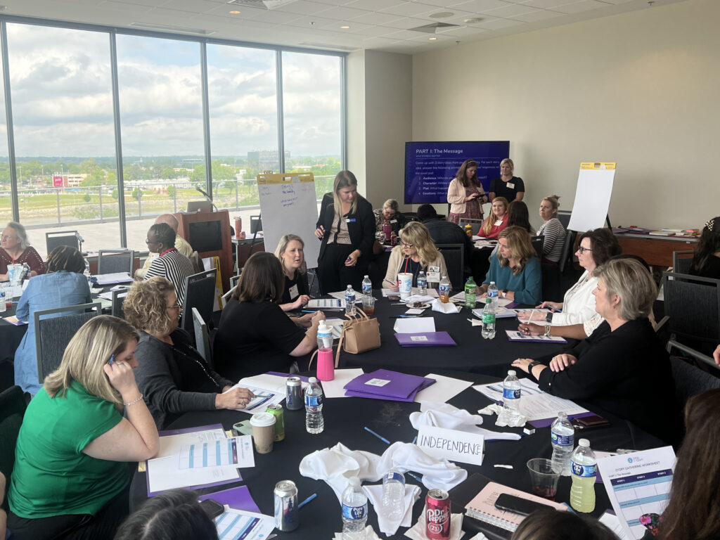 Several people are gathered around round tables in a conference room with large windows. They are participating in a group activity. The tables are covered in papers and bottled drinks, and there is a TV screen projecting a slide show. Positioned around the room are two flip charts on easels.