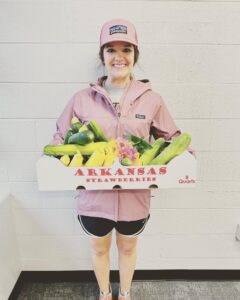 A woman in a pink hat, pink jacket, and shorts standing in a room and holding a box of vegetables grown at Batesville Preschool.
