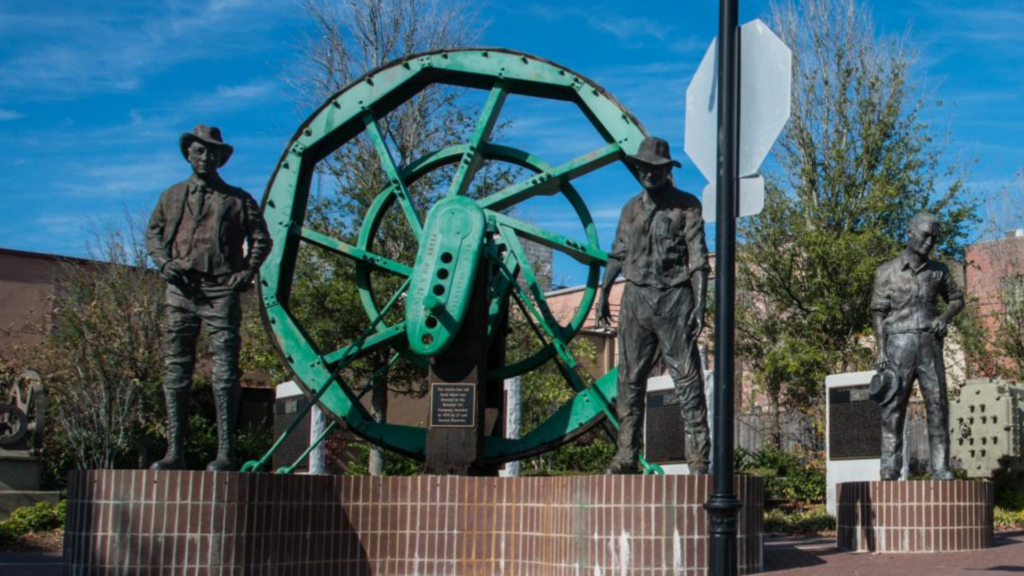 Large green wheel centerpiece surrounded by life-size bronze statues of oil workers in downtown Union County, Arkansas.