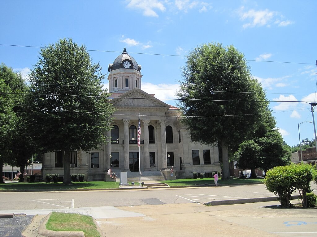 The Poinsett County Courthouse in Harrisburg, Arkansas, with a distinctive clock tower and columns, framed by tall trees on a bright summer day.